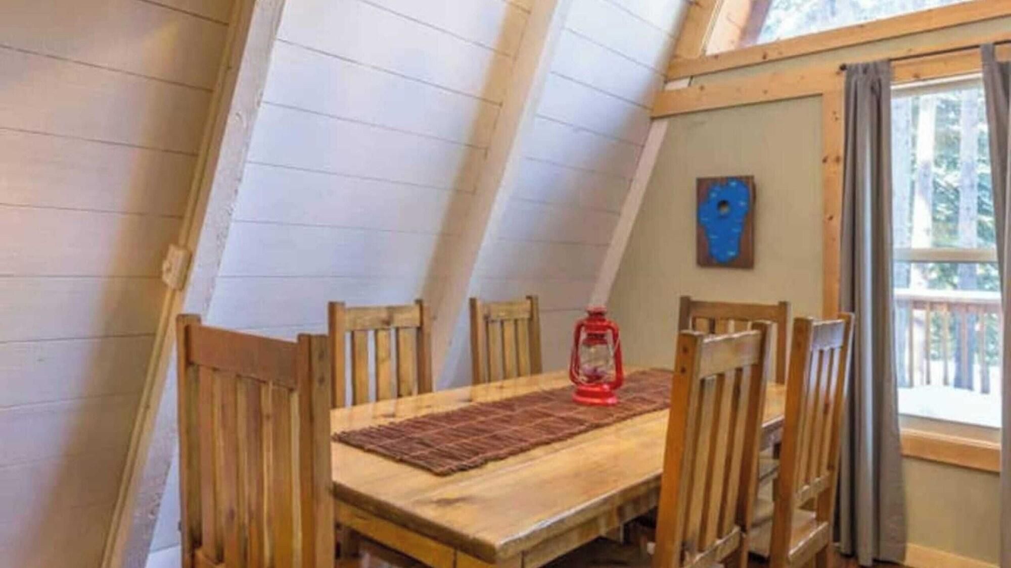 A room with a sloped ceiling, wooden walls, a table with chairs, a red lantern, and a window with curtains.