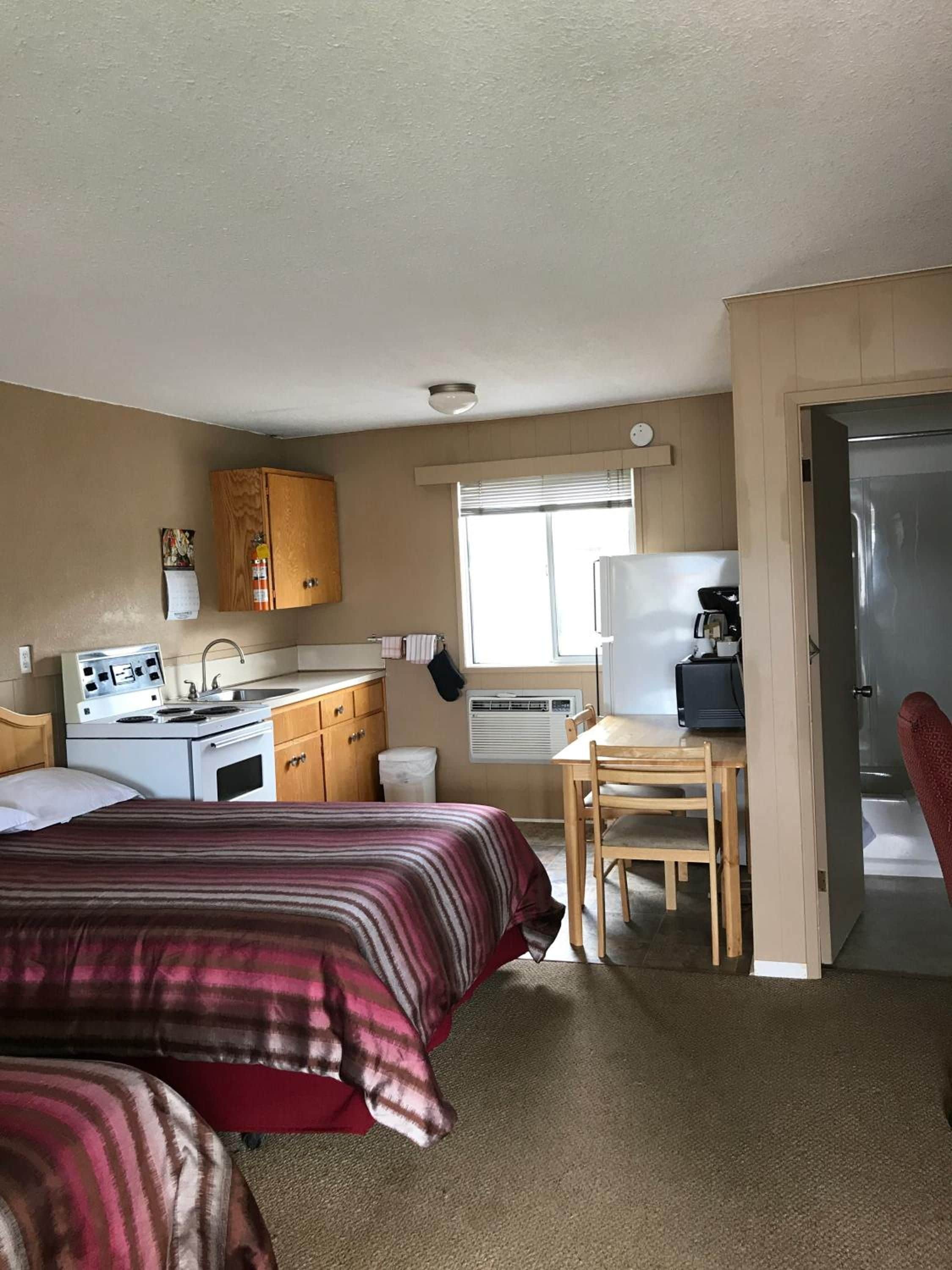 A compact room with a bed, a kitchenette, a dining area, and a door leading to another room.