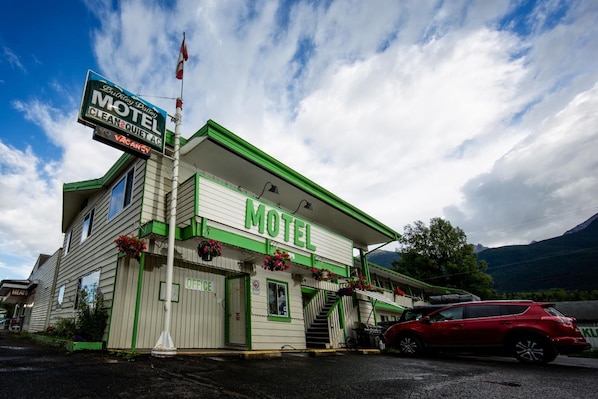 Un motel con fachada verde y blanca, una bandera en la azotea y un coche rojo estacionado al frente.