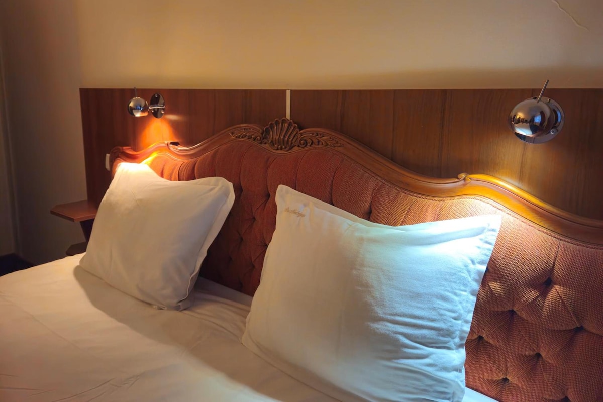A bed with white linens and two pillows, a wooden headboard with carved details, and two wall-mounted light fixtures.