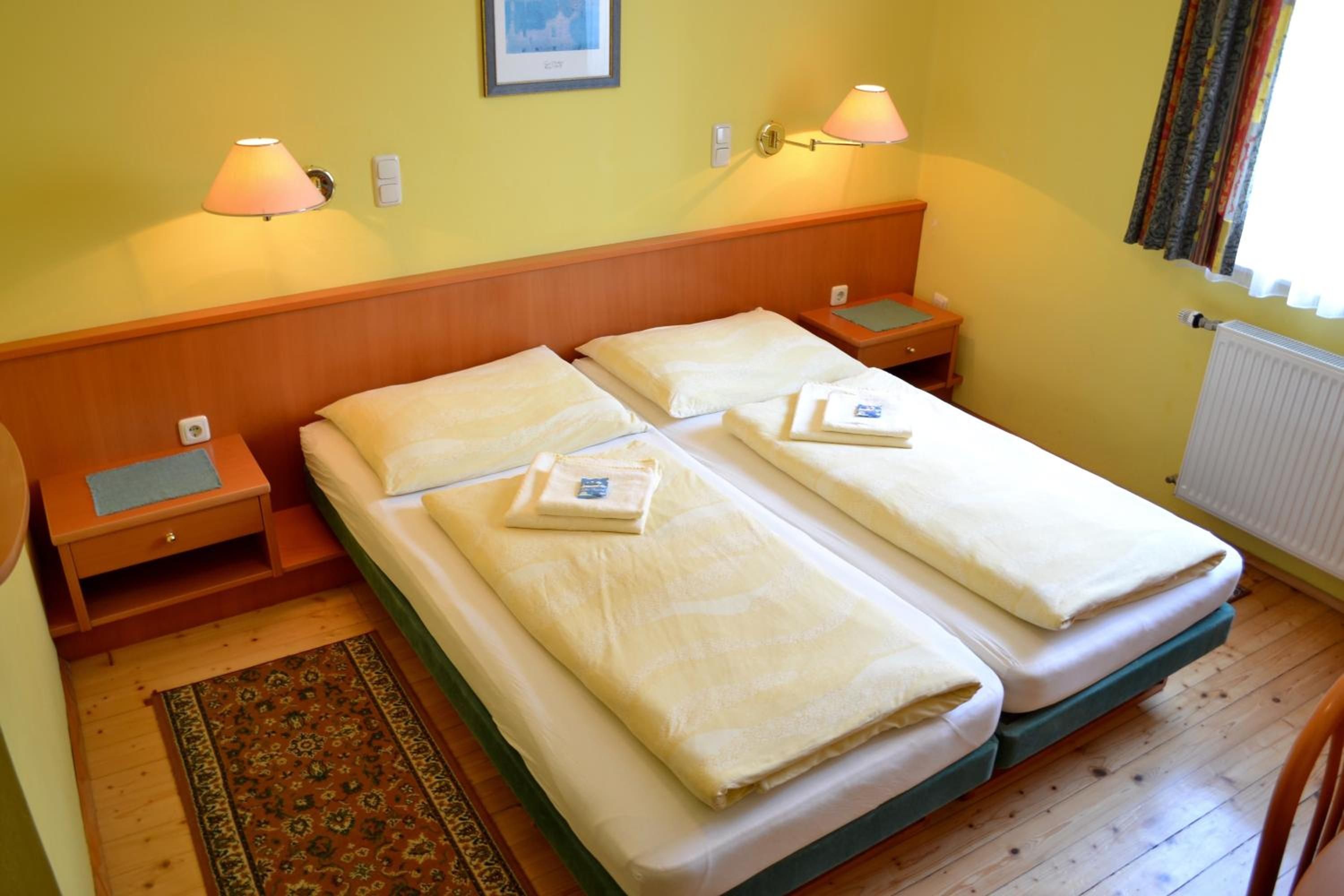 Ein Doppelbett mit zwei Kissen, ein Nachttisch mit Lampe, einer kleinen Schublade und einem gemusterten Teppich auf Holzdielen.