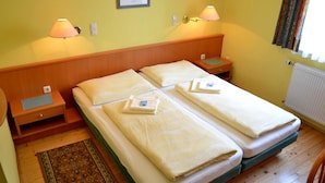 A double bed with two pillows and a folded towel, a bedside table with a lamp, a small drawer, and a patterned rug on a wooden floor. - Gasthof Redl (Oed-Öhling)