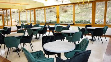 A spacious indoor area with multiple round tables and chairs, large windows offering a view of the outdoors, and a chandelier.