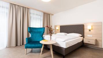 A hotel room with a bed, a blue armchair, a small table with a book, and a vase with flowers.