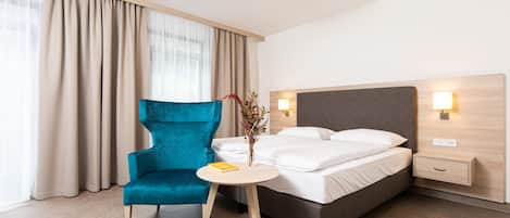 A hotel room with a bed, a blue armchair, a small table with a book, and a vase with flowers.