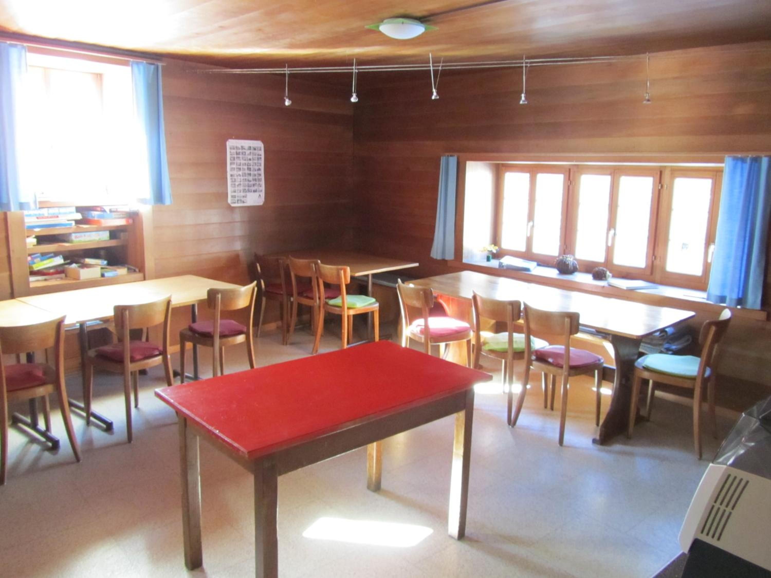 Ein Zimmer mit Holzwänden, einem roten Tisch, mehreren Stühlen, einem Bücherregal und einem Fenster mit Vorhängen.