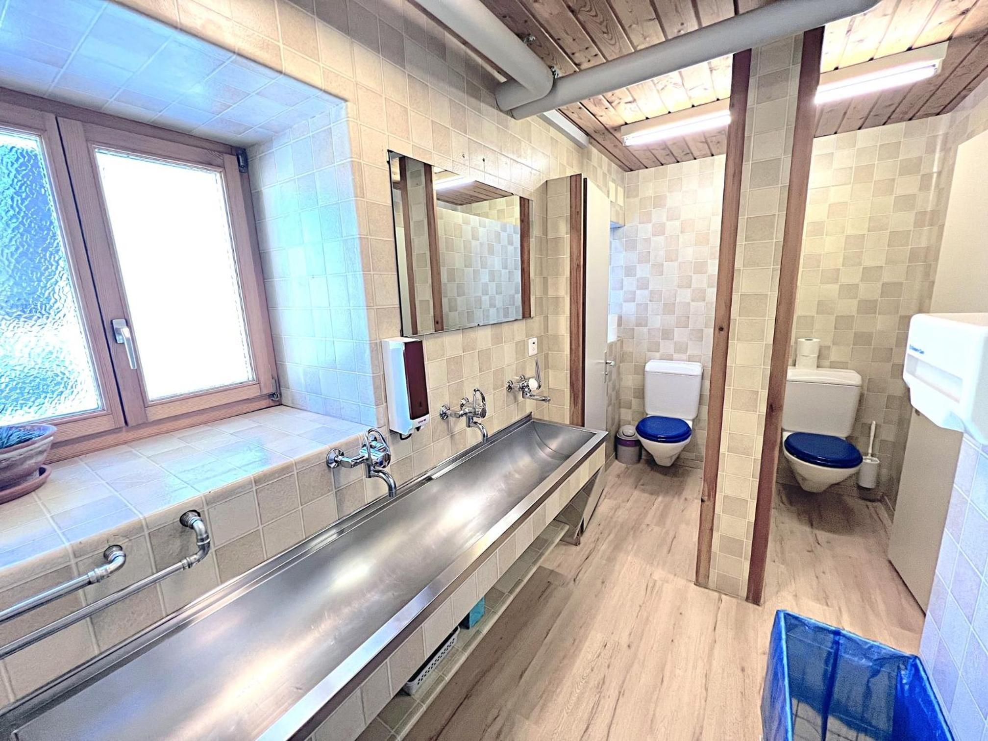 Ein modernes Badezimmer mit einem langen Edelstahlwaschbecken, zwei Toiletten und einem Fenster.