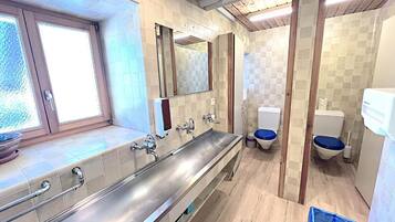 Ein modernes Badezimmer mit einem langen Edelstahlwaschbecken, zwei Toiletten und einem Fenster.