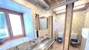 A modern bathroom with a long stainless steel sink, two toilets, and a window. - Jugendherberge Hospental (Hospental)