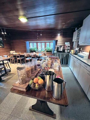 A breakfast buffet setup with a variety of food items and drinks on a wooden table, surrounded by chairs and a kitchen area. - Jugendherberge Hospental (Hospental)