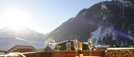 A table with a yellow tablecloth, two wine glasses, and a bottle of wine, set against a mountainous backdrop.