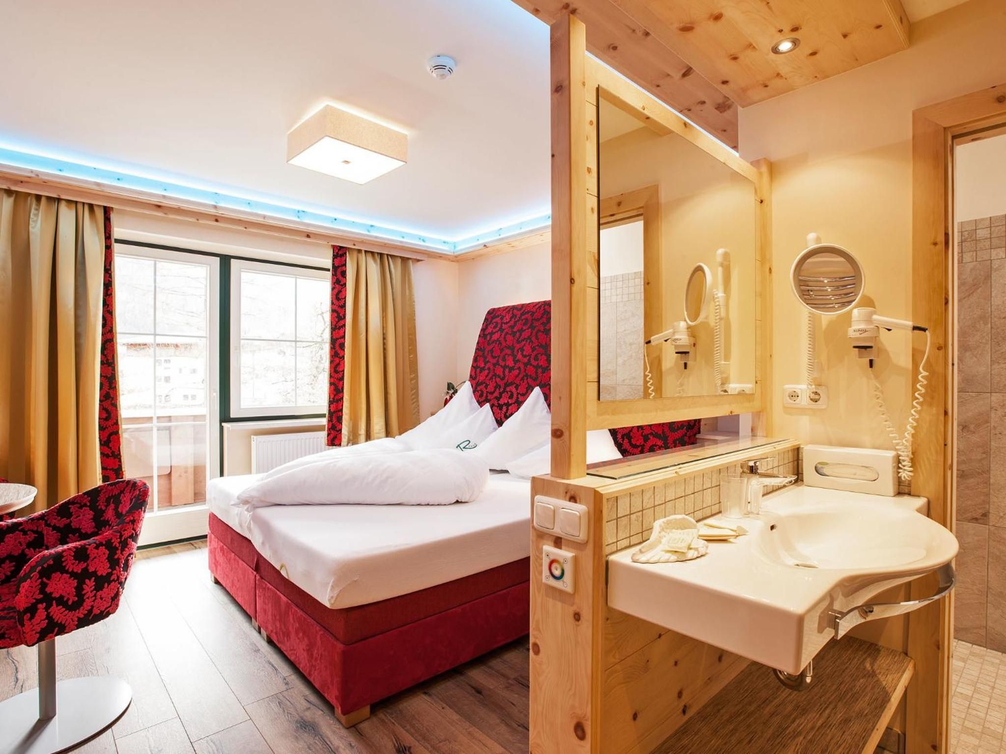 Una camera d'albergo con un letto, una poltrona rossa, un bagno con lavandino e specchio, e una finestra con tende.