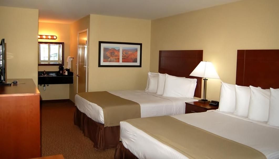 A hotel room with two beds, a desk, a mirror, a sink, and a framed picture on the wall.