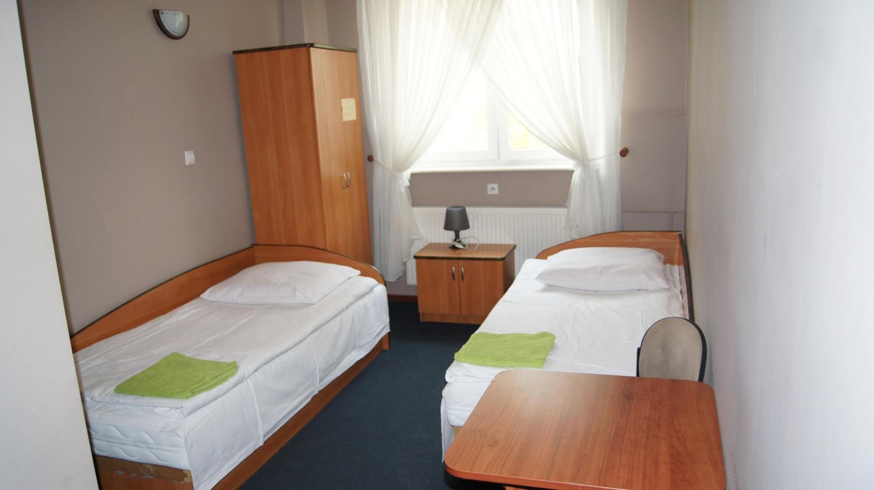 A small, compact room with two single beds, a wooden wardrobe, a small table, and a window with curtains.