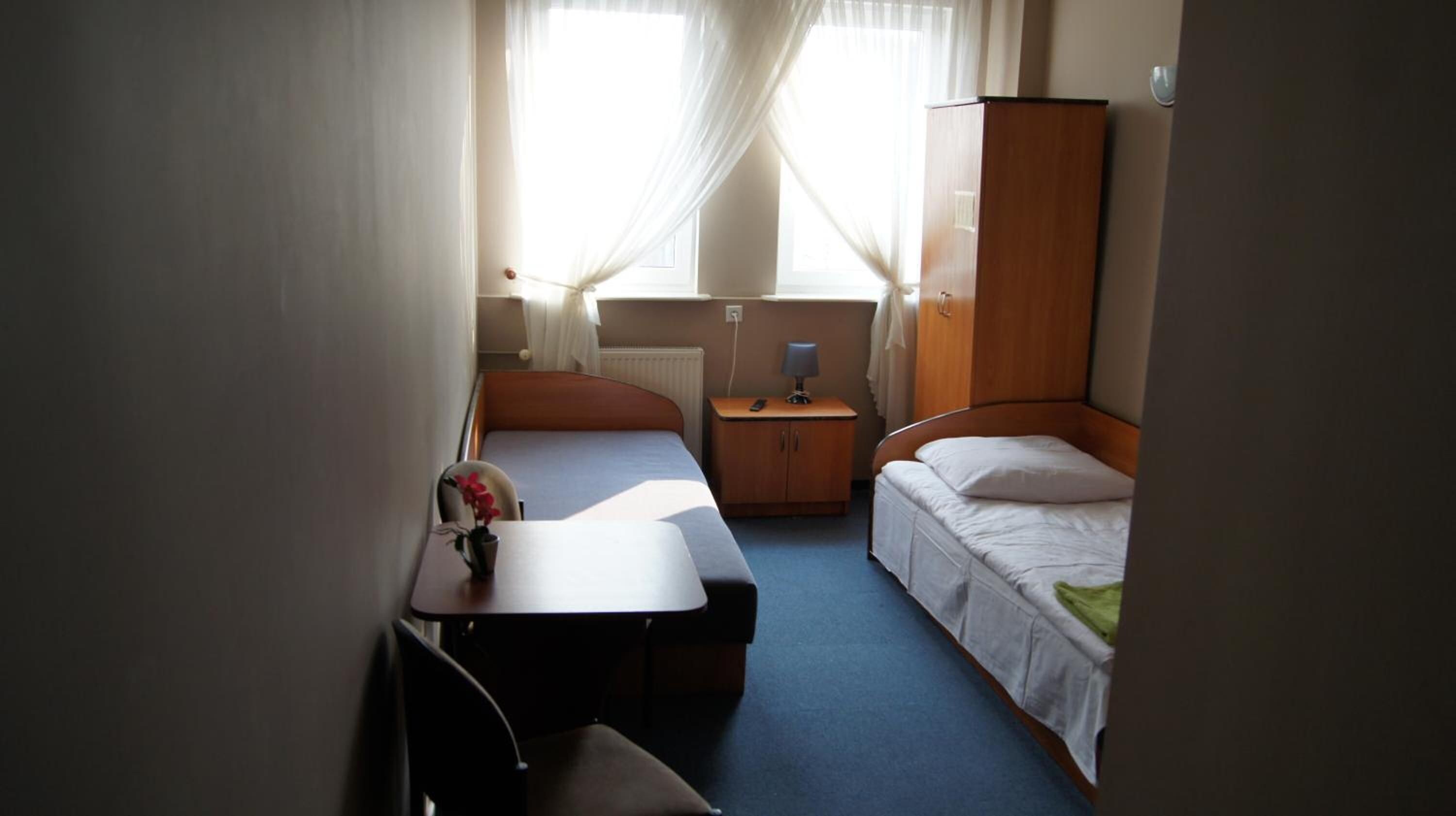 A compact hotel room with two single beds, a small table with a vase, a nightstand with a lamp, and a wardrobe.