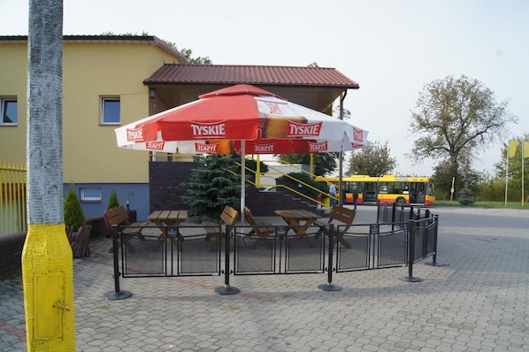 An outdoor seating area with tables and chairs under a branded umbrella, a staircase leading to a building, and a bus in the background.