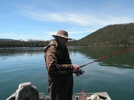 A person fishing on a boat with a calm lake and forested hills in the background.