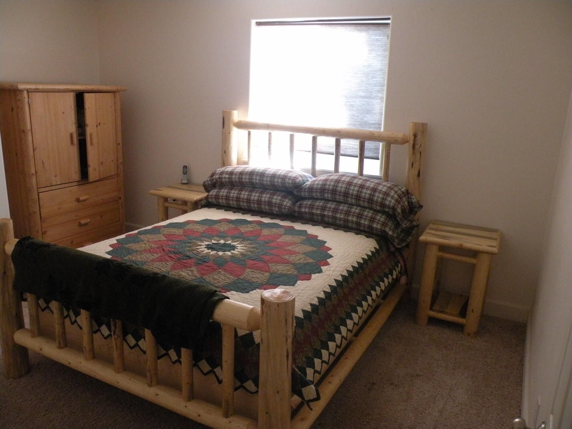 A wooden bed with a quilt, a nightstand, a wooden wardrobe, and a window with blinds.