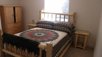 A wooden bed with a quilt, a nightstand, a wooden wardrobe, and a window with blinds.