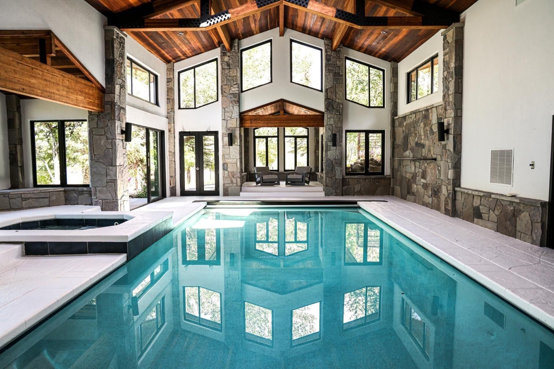 Una amplia piscina interior con paredes de piedra y vigas de madera, rodeada de grandes ventanales.