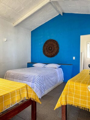A room with a bed, two tables with yellow tablecloths, a blue wall, and a woven wall hanging.