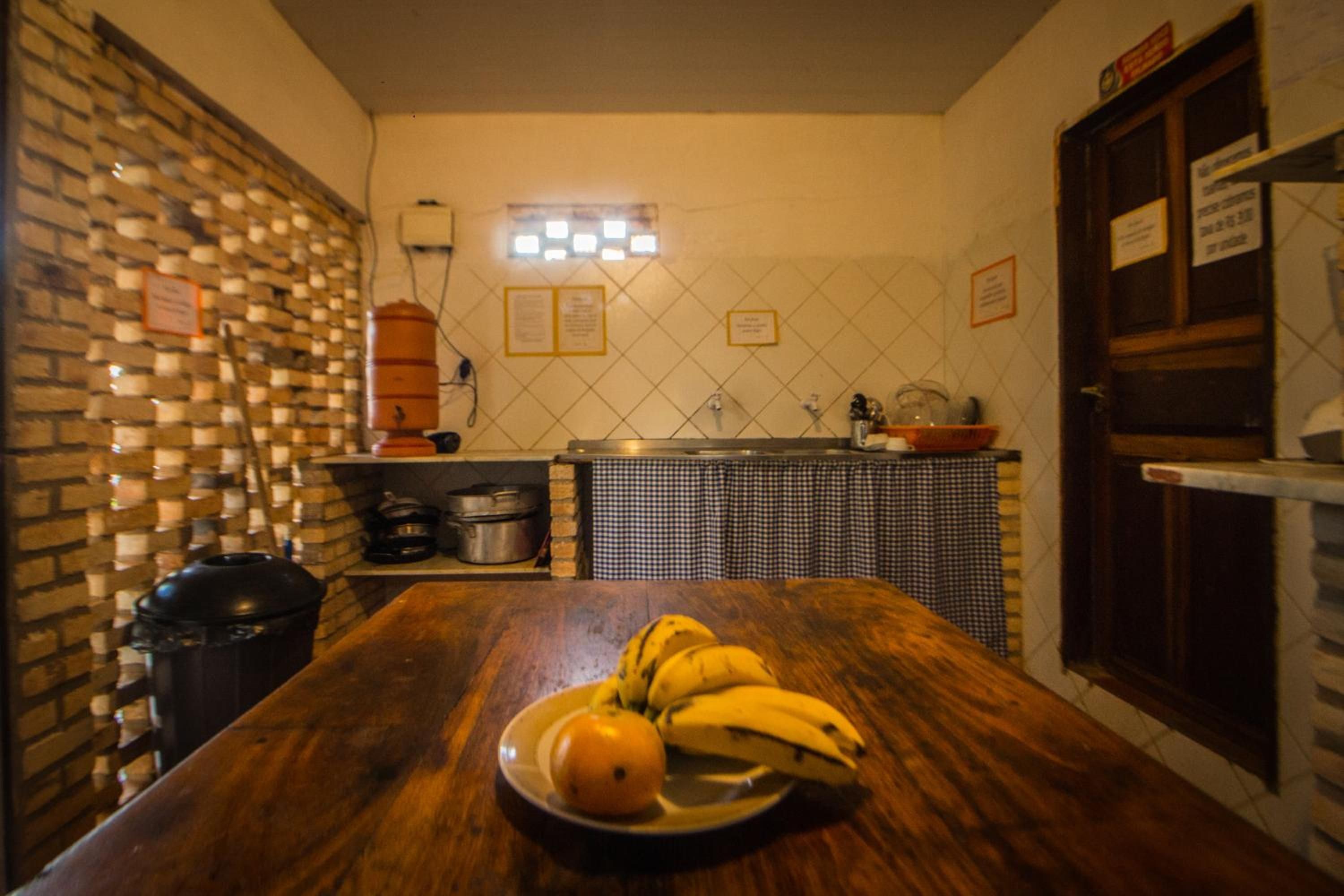 Cozinha com mesa de madeira, tigela de frutas com bananas e laranja, parede de tijolos e painel de azulejos.