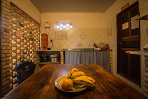 Cozinha com mesa de madeira, tigela de frutas com bananas e laranja, parede de tijolos e painel de azulejos.