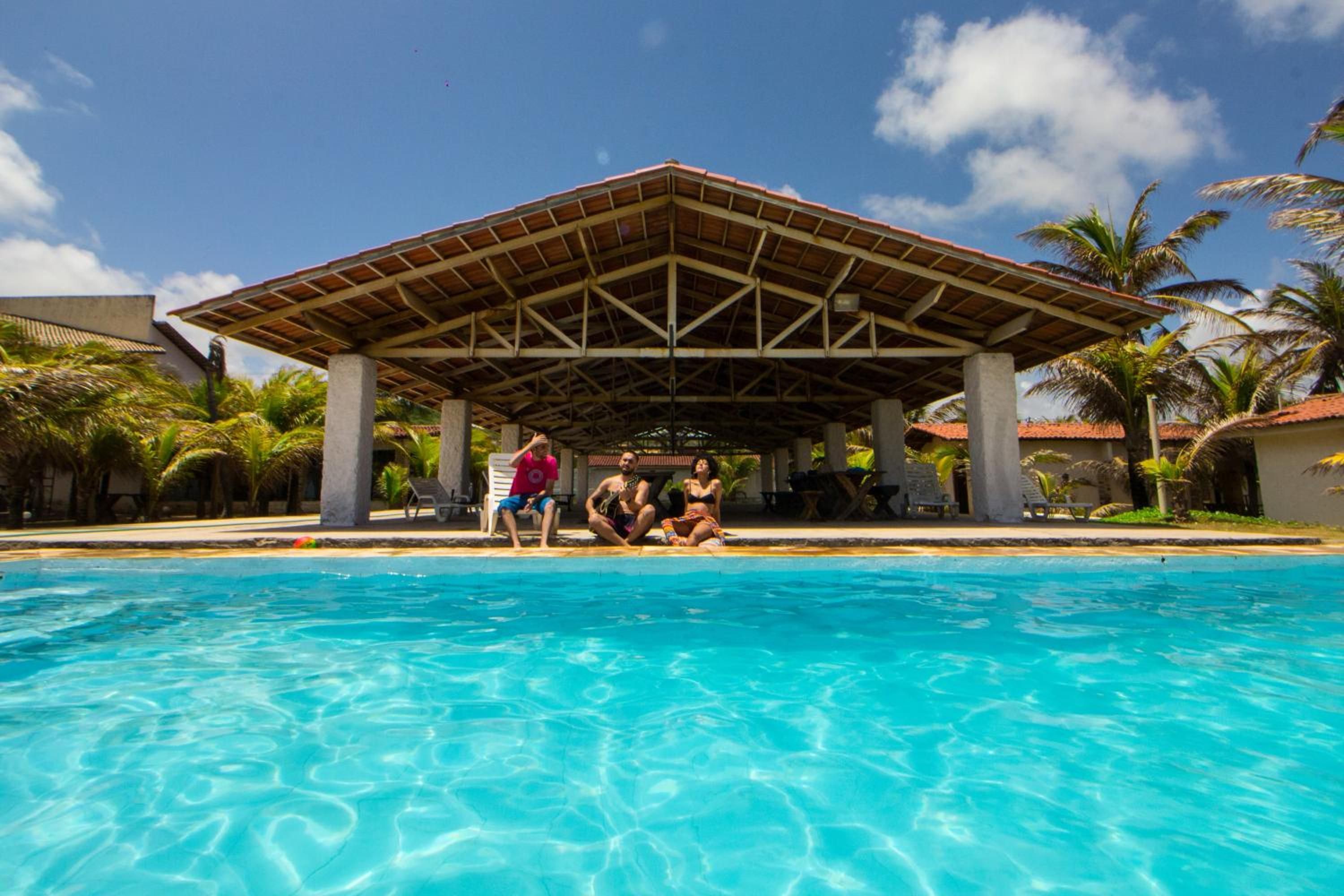 Piscina com um pavilhão coberto, palmeiras e céu azul límpido.