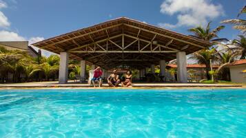 Piscina com um pavilhão coberto, palmeiras e céu azul límpido.