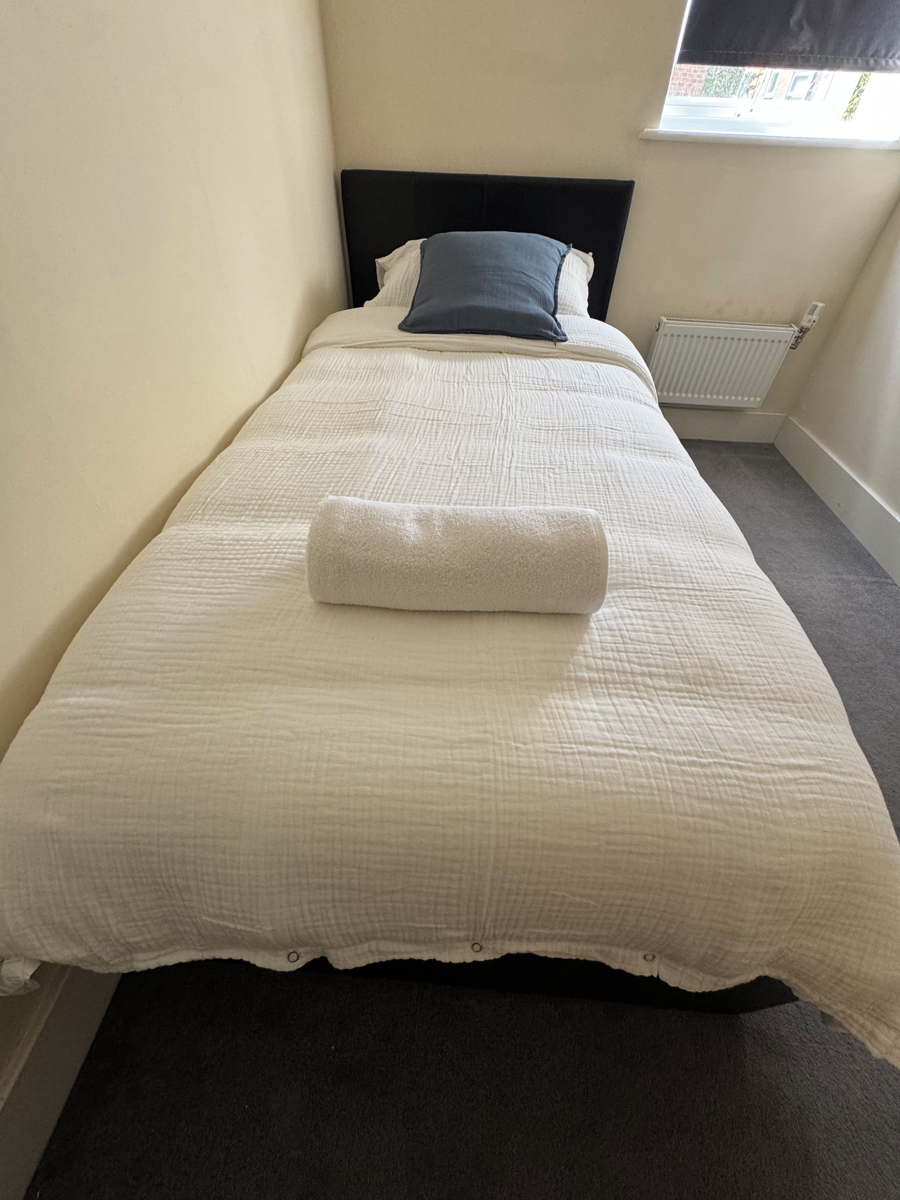 A neatly made bed with a white duvet and a rolled white towel, a dark headboard, and a window with a view of the outside.