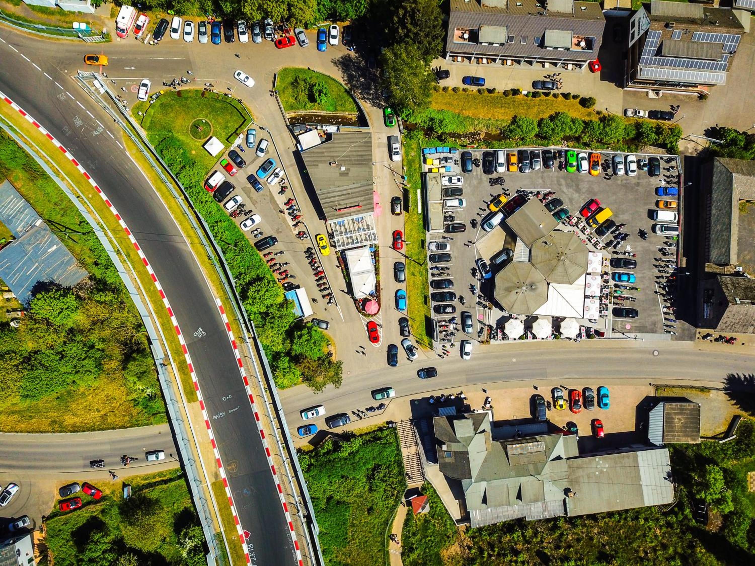 Luchtbeeld van een parkeerplaats met meerdere rijen auto's, een rotonde en omliggende gebouwen.