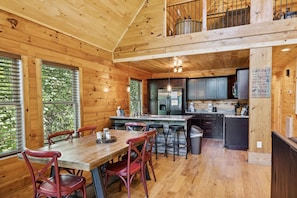 Interior de una cabaña con comedor de madera, cocina y balcón con vistas al exterior.