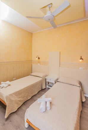 Habitación de hotel con dos camas individuales, ventilador de techo, mesita de noche con lámpara y un espejo grande en la pared.