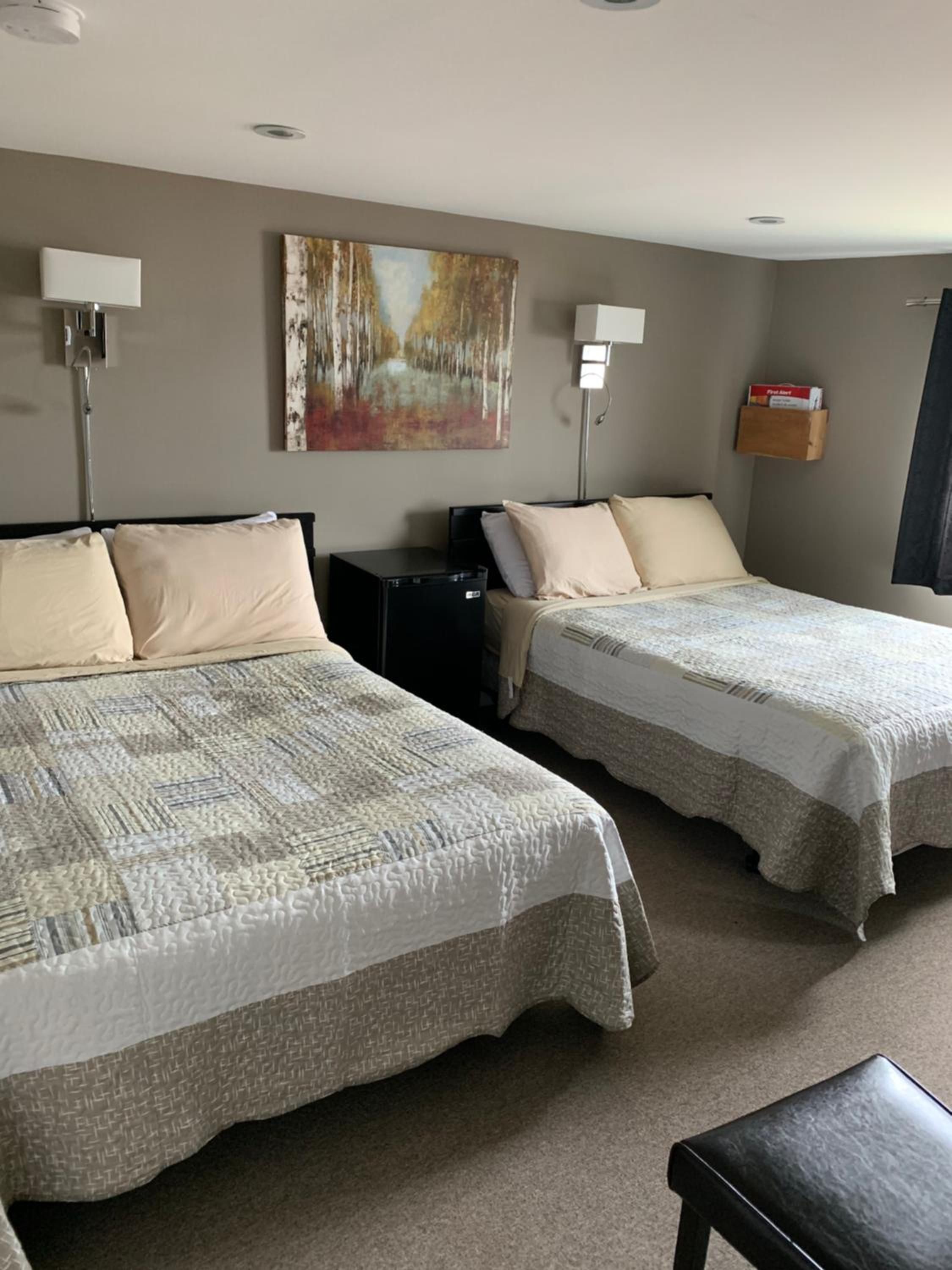 A hotel room with two beds, a desk, a lamp, a painting on the wall, and a small shelf with items.