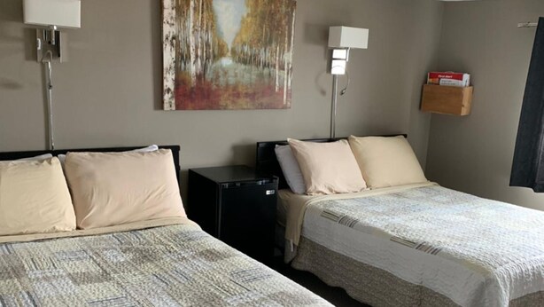 A hotel room with two beds, a desk, a lamp, a painting on the wall, and a small shelf with items. - Exploits Inn And Suites (Botwood)
