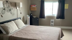A bedroom with a bed, bedside lamps, a heater, a window with curtains, and a wall decoration. - Exploits Inn And Suites (Botwood)