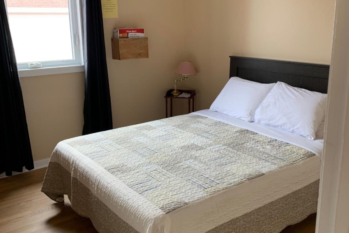 A bedroom with a bed, a window with curtains, a bedside table with a lamp, and a wall-mounted box.