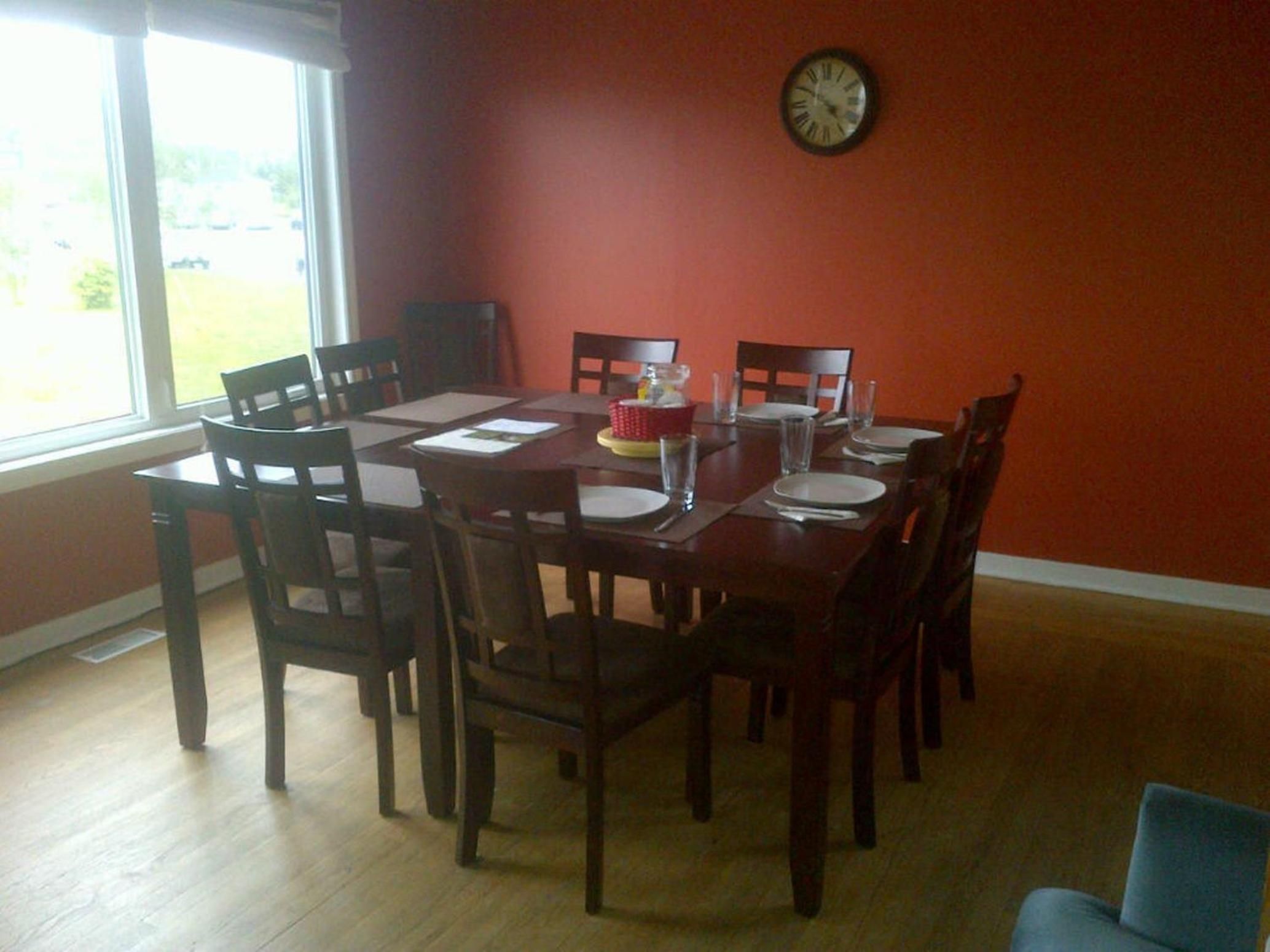 A dining area with a wooden table and chairs, a clock on the wall, and a window providing a view outside.