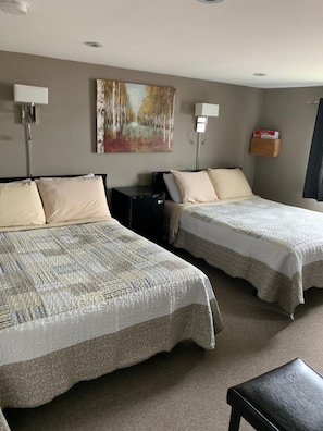 A hotel room with two beds, a desk, a lamp, a painting on the wall, and a small shelf with items.
