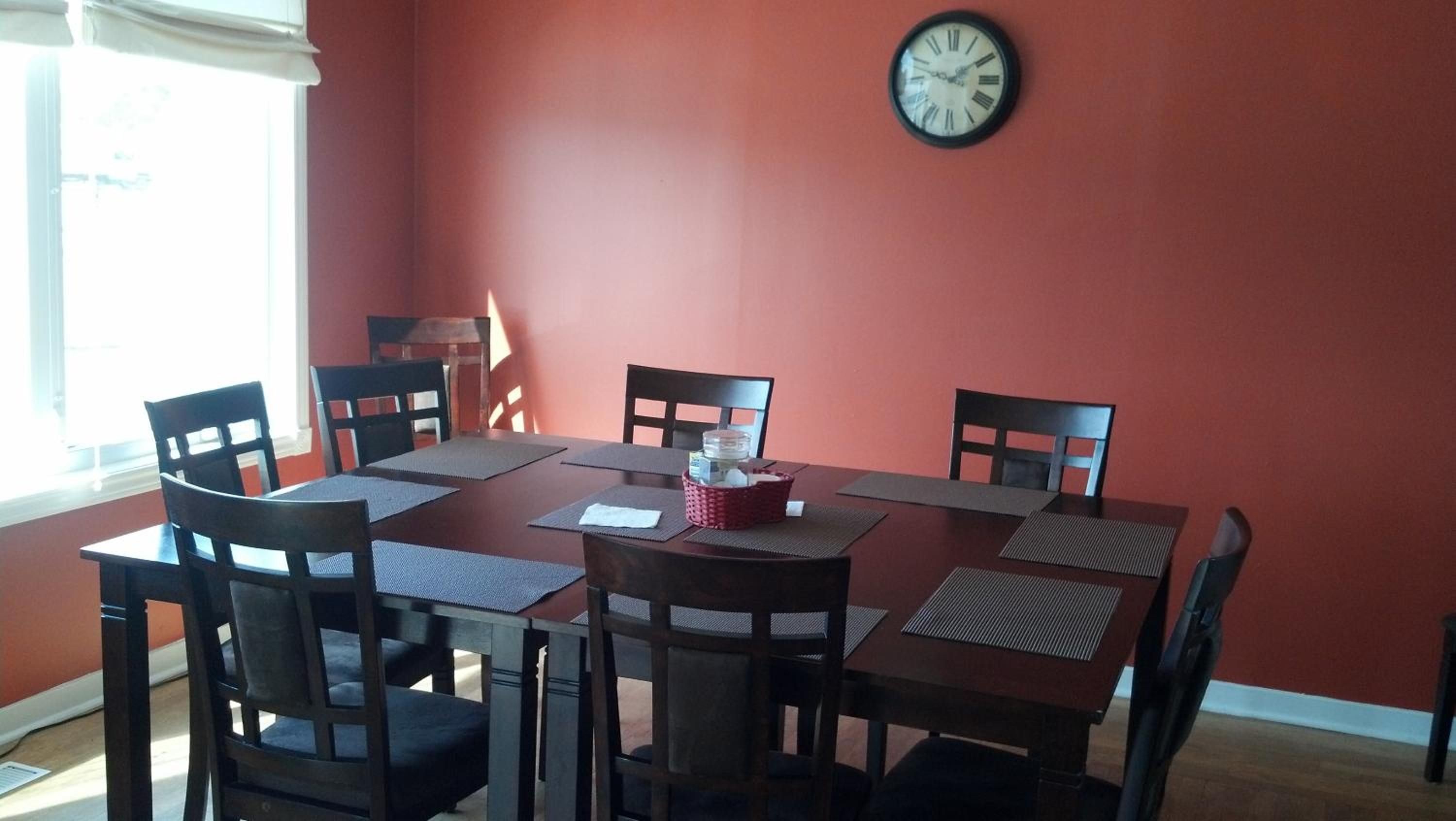 A dining area with a red wall, a round table, and six chairs. A clock is mounted on the wall.