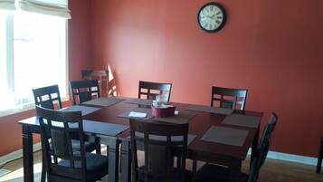 A dining area with a red wall, a round table, and six chairs. A clock is mounted on the wall.