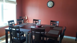A dining area with a red wall, a round table, and six chairs. A clock is mounted on the wall.