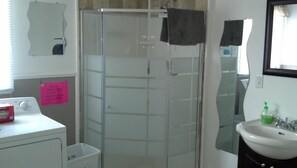 A bathroom with a glass shower enclosure, a white sink, a mirror, and a laundry machine.