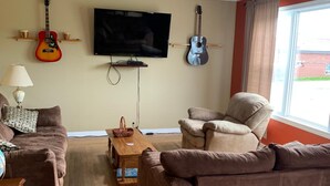 A living room with a sofa, armchair, television, guitar, and a window. - Exploits Inn And Suites (Botwood)