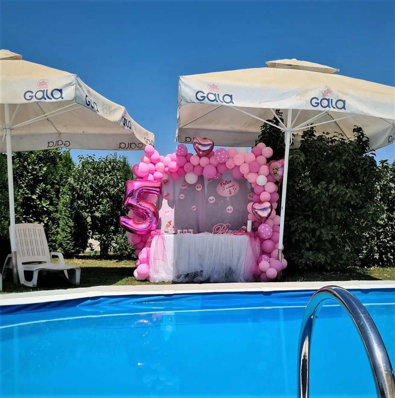 A poolside area with a pink-themed birthday setup, including a tent, balloons, and a table with a cake.