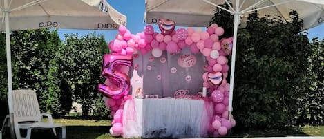 Uma área na piscina decorada para um aniversário com tema rosa, incluindo uma tenda, balões e uma mesa com bolo.