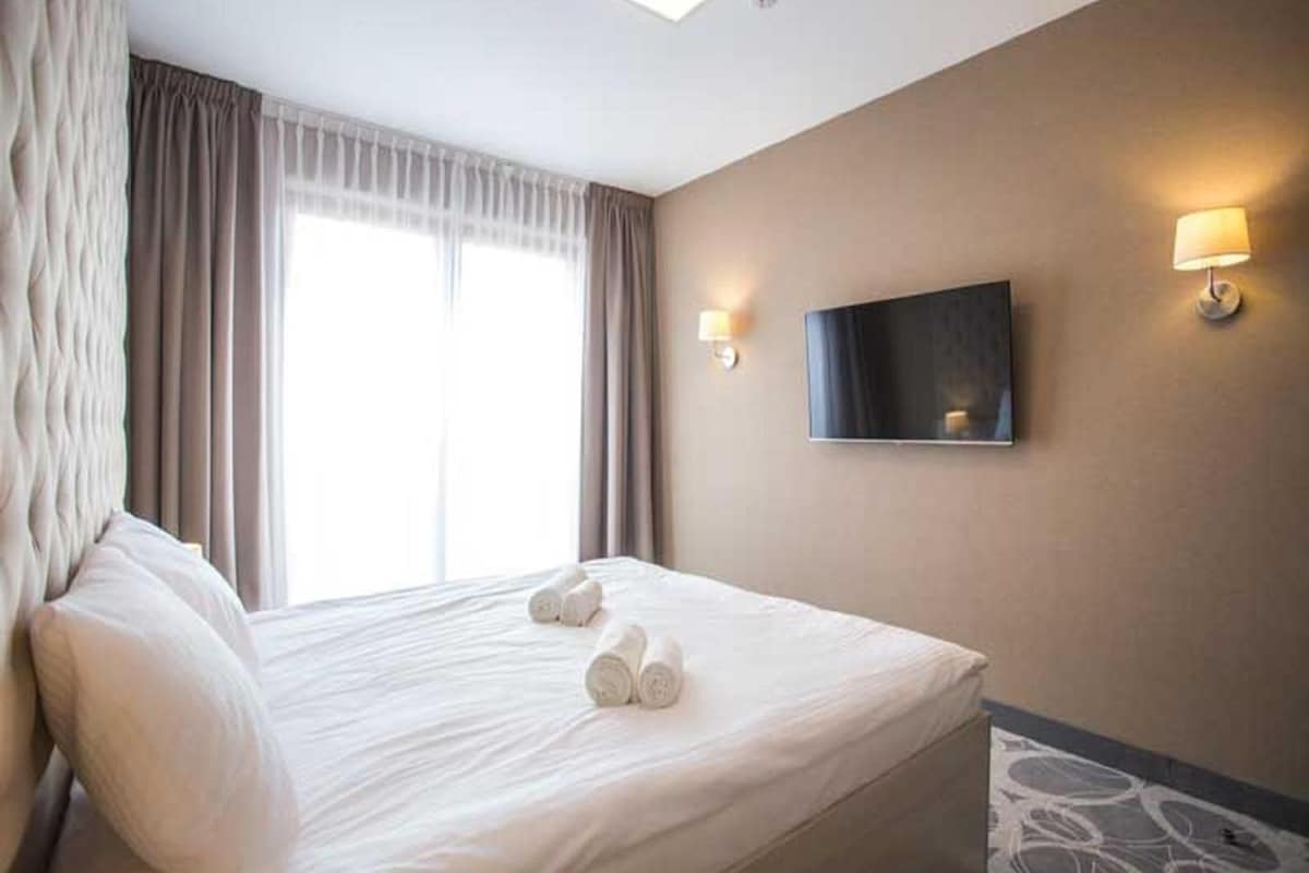 A hotel room with a large bed, curtains, a flat-screen TV, and a wall-mounted lamp.