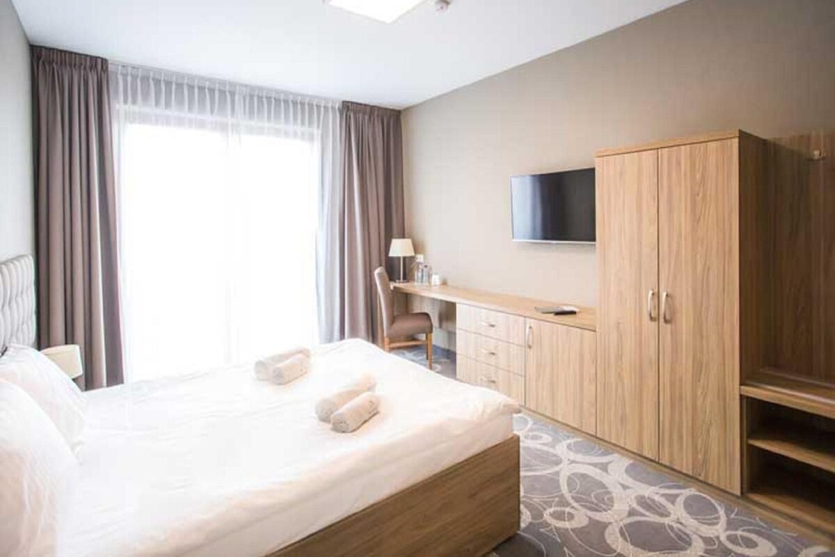 A hotel room with a large bed, a desk, a chair, a TV, and a wardrobe.