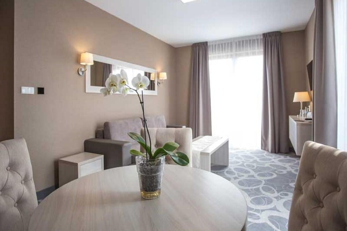 A hotel room with a round table, a sofa, a chair, a small side table, a potted plant, and a window with curtains.