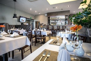 A restaurant interior with multiple dining tables, chairs, a bar area with shelves of bottles, and a television mounted on the wall.